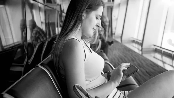 Black and white portrait of young woman with smartphone sitting in waiting room at airport terminal — Stock Photo, Image