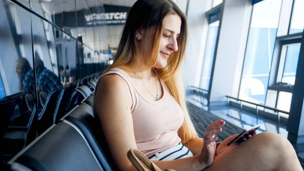 Porträt einer lächelnden jungen Frau, die auf dem Flughafen auf ihren Flug wartet und auf ihrem Smartphone in den sozialen Medien surft — Stockfoto