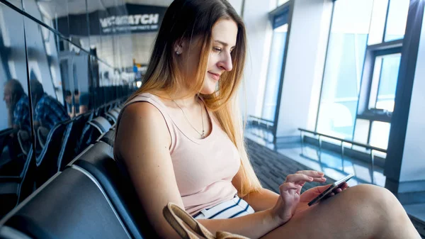 Closeup portrait of beautiful blonde woman sitting in airport waiting room and browsing internet on smartphone — Stock Photo, Image