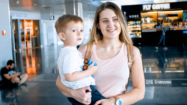 幼児の少年を抱き、ショッピングモールや空港ターミナルを歩く美しい笑顔の女性の肖像画 — ストック写真