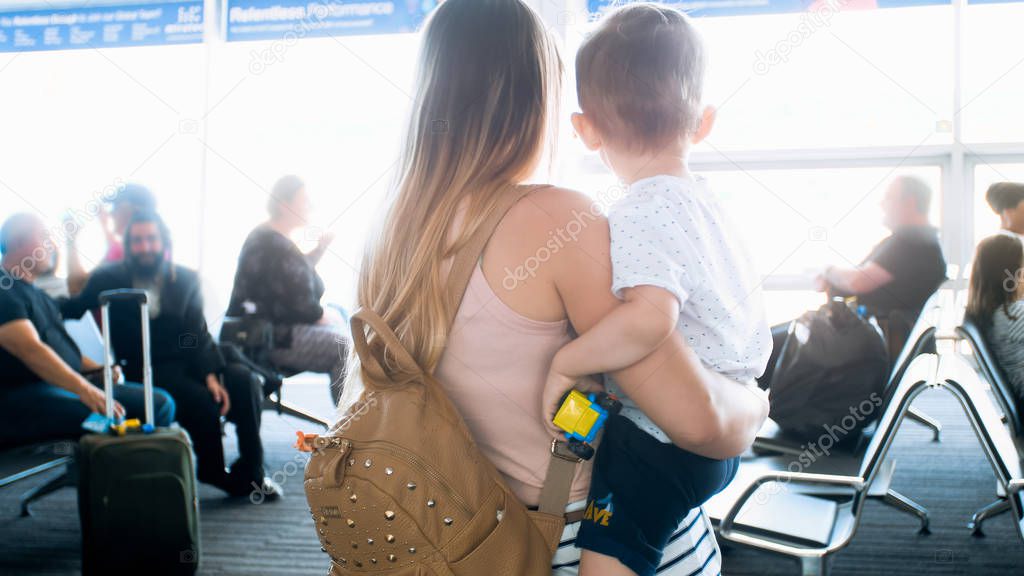Rear view image of young woman holding her toddler son and looking through big window in airport terminal