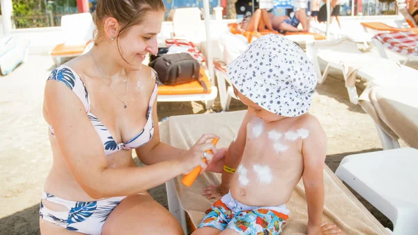 Young mother using sunblock spray to protect her child from UV sun light on beach — Stock Photo, Image