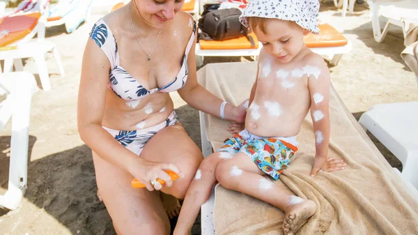 Young smiling woman spraying sunblock lotion over her little son sitting on sunbed at sea beach — Stock Photo, Image