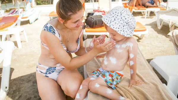 Young woman applying sunscreen lotion on her little child sitting on sunbed at sea beach — Stock Photo, Image
