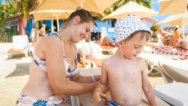 Schöne junge Mutter sitzt mit ihrem Kind auf der Sonnenbank am Strand und cremt sich ein — Stockfoto