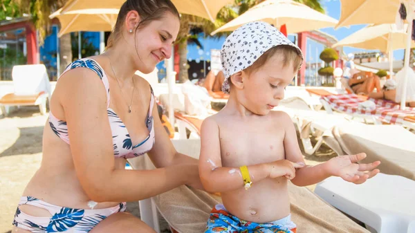 Beautiful young mother using sun protection lotion on her childs sking before sunbathing at sea beach — Stock Photo, Image