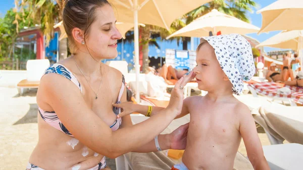 Schöne junge Mutter sitzt mit ihrem Kind auf der Sonnenbank am Strand und cremt sich ein — Stockfoto