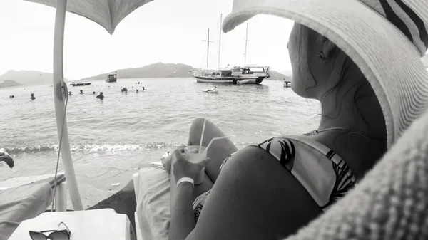 Foto de vista trasera en blanco y negro de una mujer joven con sombrero grande relajándose en la playa con cóctel —  Fotos de Stock