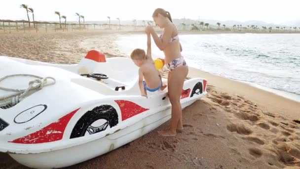 4k filmagem de jovem mãe ajudando seu filhinho escalando em catamarã de pé na praia do mar — Vídeo de Stock