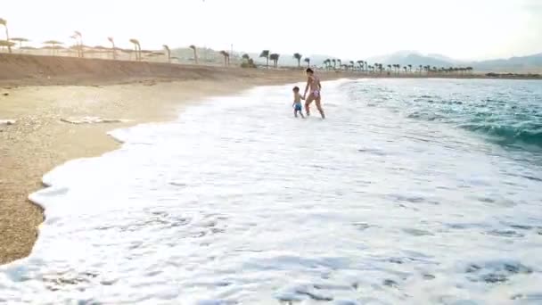 4k video van gelukkige jonge moeder met haar kleine zoon wandelen en rennen in de zee golven bij zonsondergang. Familie ontspannen op het strand op zomer vakantie — Stockvideo