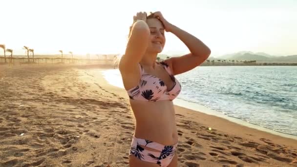 4k video o usmívající se mladé ženě v bikinách pózách na pláži při západu slunce. Mladá dívka stojící na moři a dívá se do kamery — Stock video