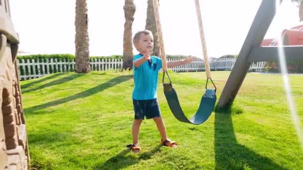 4k metraje de niño alegre jugando en el patio de recreo con columpio en el soleado día de verano — Vídeos de Stock