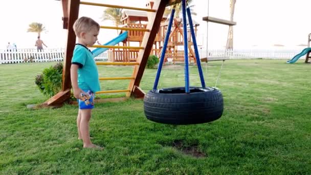 4k видео веселого мальчика, играющего на детской площадке с качелями в солнечный летний день — стоковое видео
