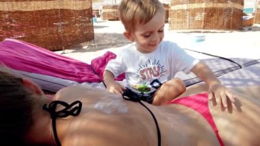 Deniz sahilinde şezlongda yatan genç anneye güneş kremi uygulayan küçük yürümeye başlayan çocuğun 4k videosu