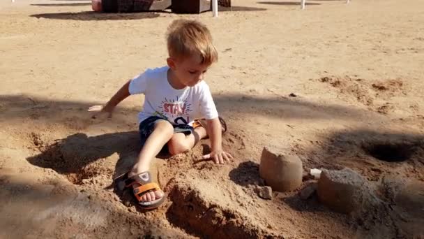 4k-beelden van kleine peuter jongen zittend op het strand van de zee en gebouw kasteel van natte zand — Stockvideo