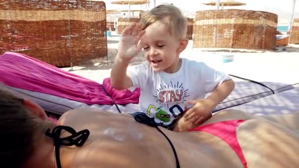 4k vídeo of cute little boy applying UV protection lotion on mothers back lying on lounger at sea beach — Vídeo de Stock