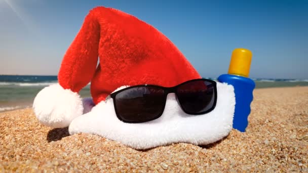 4k video di Babbo Natale cappello, occhiali da sole e crema solare lozione sdraiato sulla spiaggia sabbiosa del mare. Concetto di celebrare le vacanze invernali in vacanza estiva — Video Stock