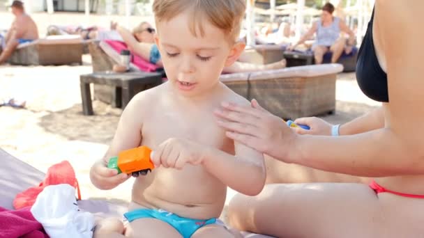 4k Nahaufnahme Video einer jungen Frau, die mit ihrem kleinen Sohn auf einer Sonnenliege am Strand sitzt. Mutter cremt ihr Kind mit Sonnencreme ein — Stockvideo