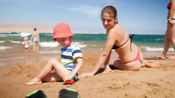 4K αργή κίνηση βίντεο του χαρούμενο μικρό αγόρι με χαμογελαστή μητέρα παίζει στην αμμώδη παραλία την ηλιόλουστη μέρα του καλοκαιριού — Αρχείο Βίντεο
