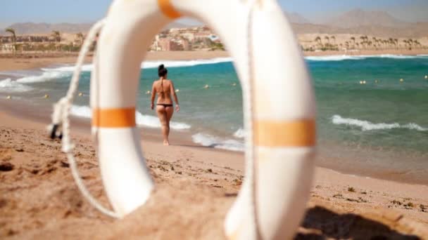 Imágenes de 4k de una joven sexy en mini bikini caminando por la playa de arena en un día ventoso. cámara mirando a través de salvavidas boya anillo — Vídeos de Stock