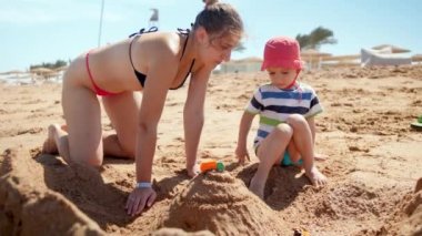 Kumlu deniz plajında dinlenirken onun bebek oğlu ile oynarken genç anne 4k yavaş hareket video