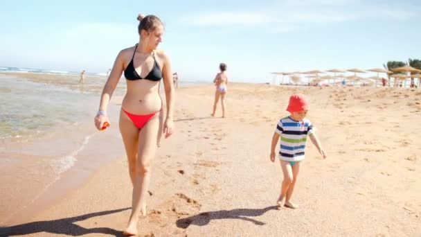 4k video van mooie jonge vrouw in bikini wandelen met haar peuter zoon op het zandstrand van de zee — Stockvideo