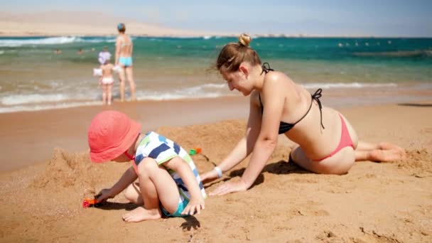 4k відео молодої матері зі своїм сином, який грає з іграшковою машиною на морському пляжі. Сімейний відпочинок під час літніх пляжних канікул — стокове відео