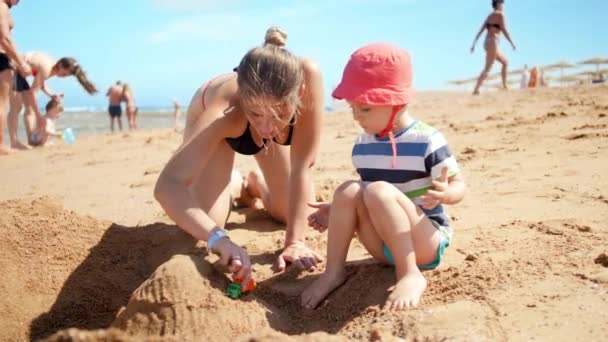 4k视频的年轻母亲与她的孩子儿子玩玩具车在海边海滩。夏季海滩度假期间的家庭放松 — 图库视频影像