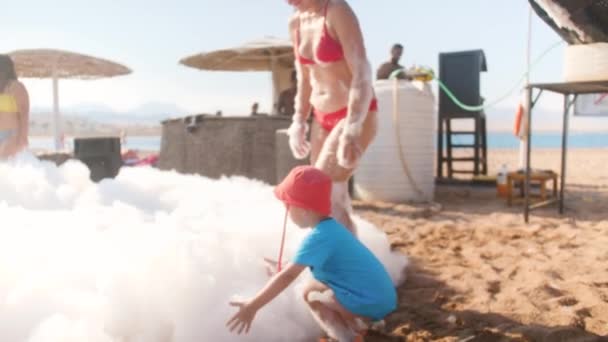 4k видео маленького мальчика с матерью, играющей с мыльной пеной на пляже во время танцев и веселых пляжных дискотек — стоковое видео