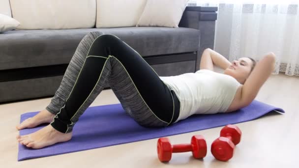 Vídeo de jovem mulher em leggings deitado no tapete de fitness na sala de estar e fazendo abdominais crunches — Vídeo de Stock