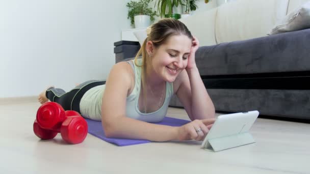 4k video de hermosa mujer joven sonriente acostada en la alfombra de fitness y el uso de la tableta digital. Chica viendo clases de fitness en gadget — Vídeo de stock