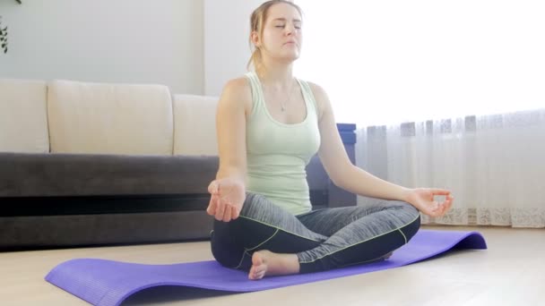 Dolly 4k Filmmaterial von schönen jungen Frau sitzt Lotus Yoga-Position auf Fitnessmatte im Wohnzimmer und meditiert — Stockvideo