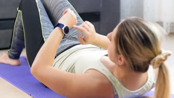 Video de una joven tumbada en una esterilla de yoga y colocando un rastreador de fitness en su muñeca antes de hacer abdominales crujidos — Vídeos de Stock