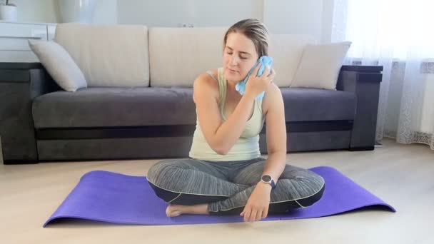 Imágenes en cámara lenta de una mujer joven agotada sentada en una esterilla de fitness y limpiando el sudor con una toalla de su frente después de hacer ejercicio — Vídeos de Stock