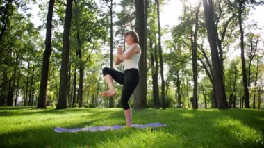 Parkta çim yoga ve fitness 4k yavaş hareket video. Orta yaşlı gülümseyen kadın pratik ve ormanda güneşli bir günde egzersizyapıyor. Kadın onun zihinsel ve fiziksel sağlığı bakımı