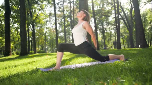 Filmagem 4k de mulher sorridente feliz que se estende no tapete de fitness no parque. Senhora de meia idade fazendo exercícios de ioga na floresta . — Vídeo de Stock