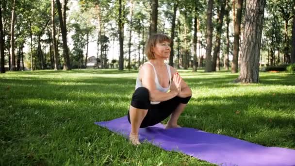 4k lassú mozgás videó a 40-es években nő gyakorló jóga füvön parkban. Felnőtt hölgy fitness gyakorlatokat végez a matracon az erdőben. — Stock videók