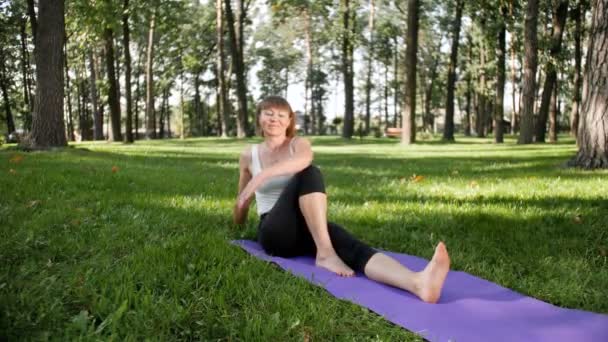 4k video en cámara lenta de una hermosa mujer de mediana edad sonriente haciendo ejercicios de yoga y estirándose en el parque. Es muy útil para la salud mental y física. Gente relajándose en la naturaleza — Vídeo de stock