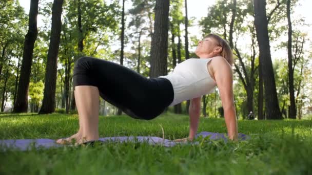 4k video en cámara lenta de una mujer sonriente de mediana edad practicando yoga y meditando en el parque en el soleado día de verano — Vídeo de stock