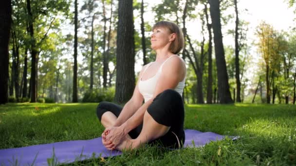 4k slow motion video of 40s woman practicing yoga on grass at park. Взрослая женщина делает упражнения на коврике в лесу . — стоковое видео