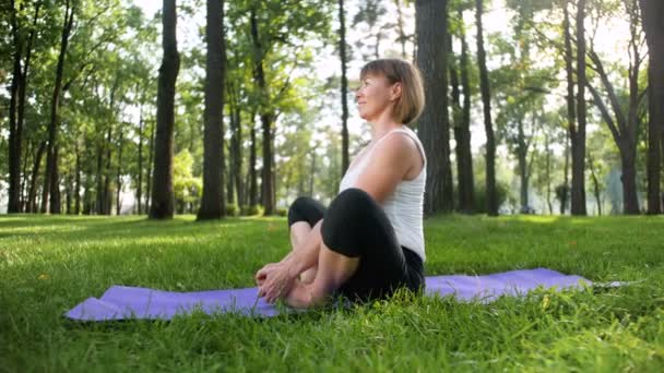 Imágenes de 4k de la feliz mujer sonriente estirándose en la alfombra de fitness en el parque. Señora de mediana edad haciendo ejercicios de yoga en el bosque . — Vídeos de Stock