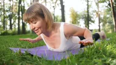 Yerel parkta mat fitness egzersizleri yapan 40 yaşındaki kadının 4k görüntüleri. Doğada zihinsel ve fiziksel sağlıklarıyla ilgi çeken insanlar