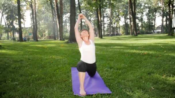 4k vídeo em câmera lenta de ioga e fitness na grama no parque. Mulher sorrindo de meia-idade praticando e fazendo exercícios no dia ensolarado na floresta. Mulher cuidando de sua saúde mental e física — Vídeo de Stock