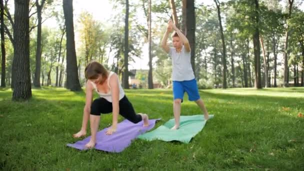 Vídeo en cámara lenta de 4k de una mujer de mediana edad enseñando a un adolescente en clase de yoga. Mujer y niño haciendo ejercicios de fitness en el parque — Vídeos de Stock