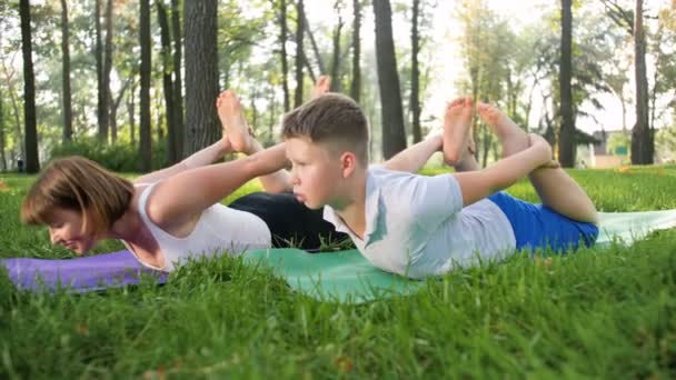 4k кадров подростка, практикующего йогу с матерью средних лет на траве в парке. Семья занимается фитнесом и растягивается в лесу — стоковое видео