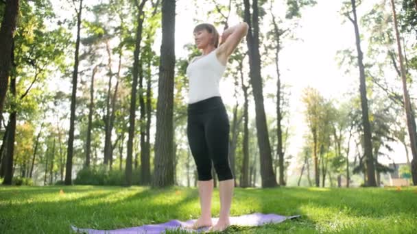 4k lassú mozgás videó középkorú mosolygó nő gyakorló jóga és meditál parkban napos nyári nap — Stock videók