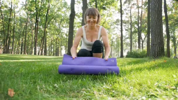 Vídeo em câmera lenta do treinador de fitness feminino desrolando tapete de ioga na grama verde no parque. Fazendo exercícios esportivos e alongamento na floresta — Vídeo de Stock