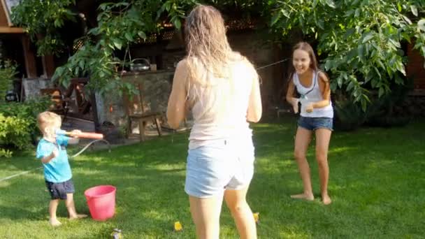 Pomalý film z veselých mladých rodin s dětmi, které si hrají s vodními pistolemi a s vodní hadičkou na zahradě v horkém letním dni — Stock video