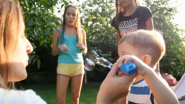 4k повільне відео про щасливу сім'ю з дітьми, що дме мильні бульбашки в саду на задньому дворі в теплий сонячний день — стокове відео