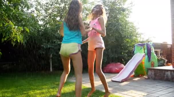 Parlak güneşli bir günde bahçe hortumundan su damlacıkları altında eğlenen neşeli genç kızların yavaş çekim videosu — Stok video